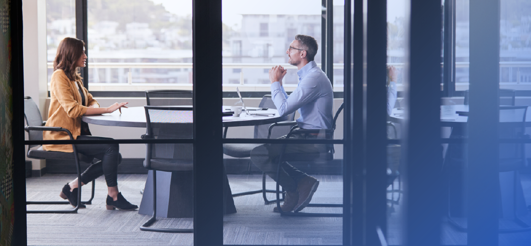 Two colleagues talking across a table in a glass-walled office meeting room
